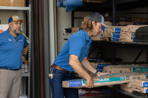 Roofing team member pulling shingle bundles from warehouse shelves while a colleague looks on