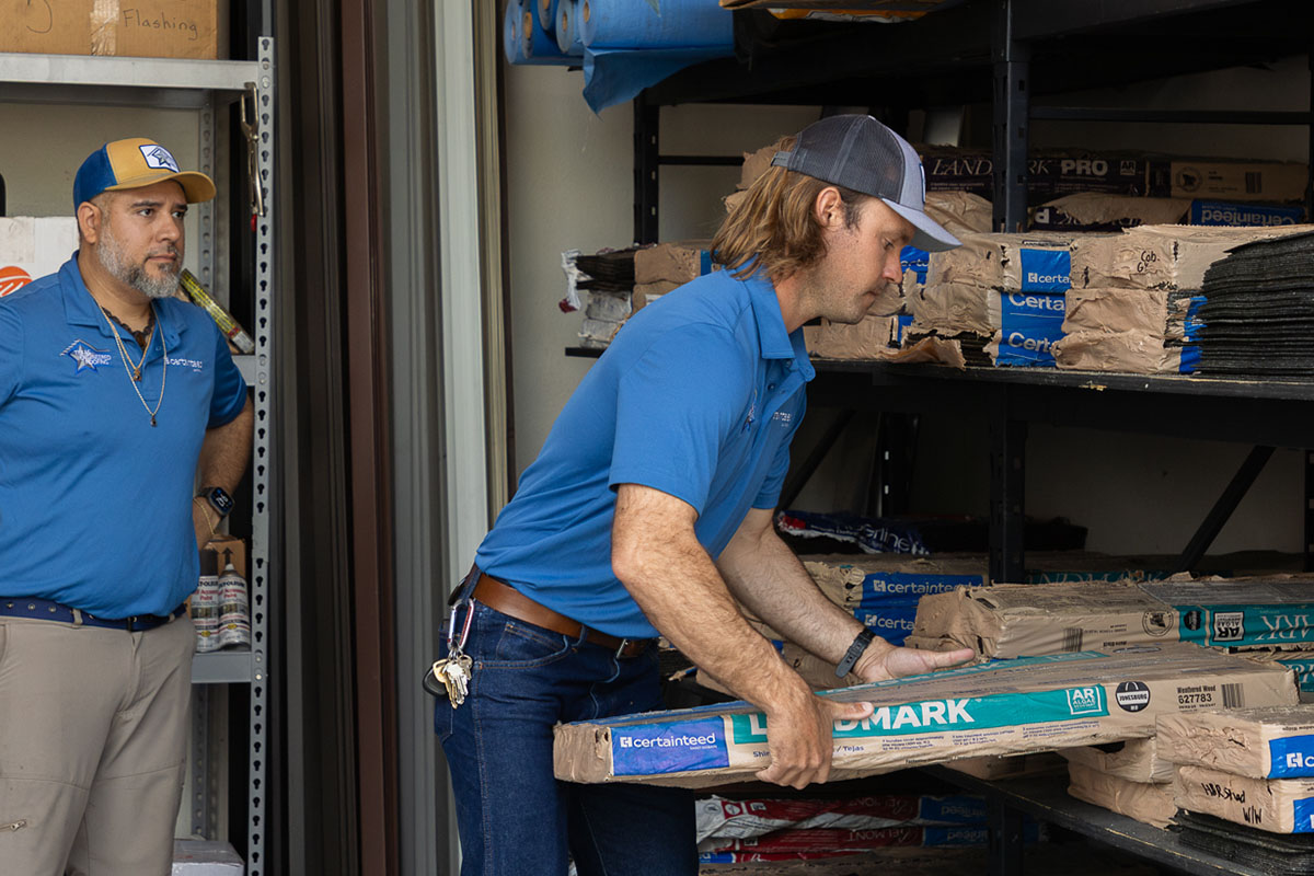 Roofing team member pulling shingle bundles from warehouse shelves while a colleague looks on