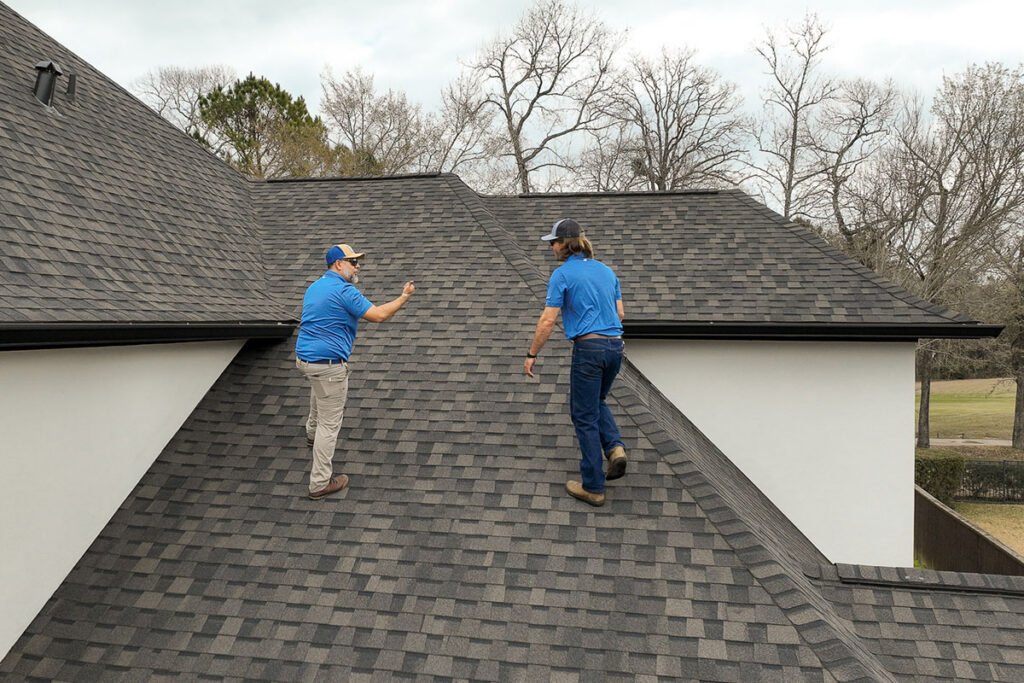 Two roof inspectors in blue shirts walking the ridge of a gray shingle roof during an inspection