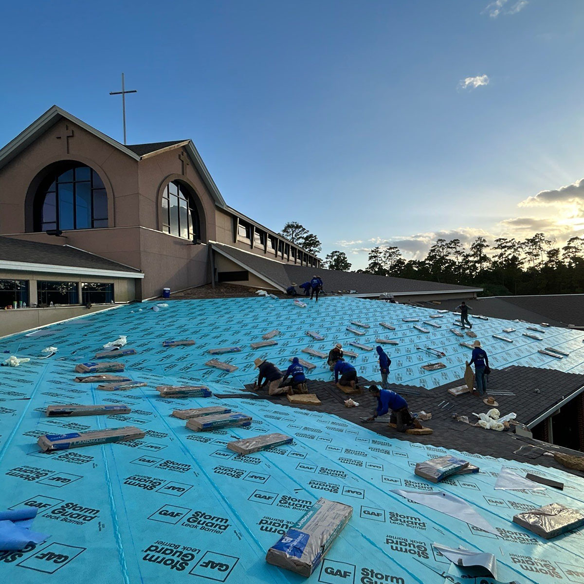 Roofing crew installing new roofing over blue underlayment on a large church roof at sunset