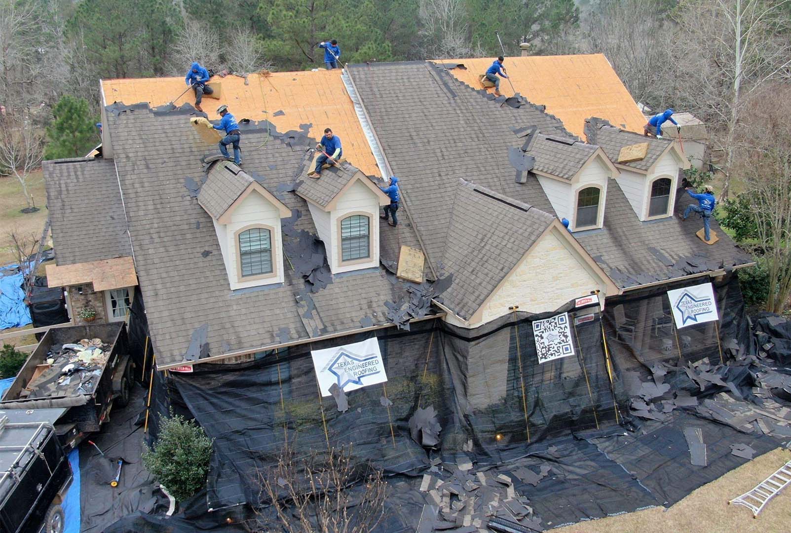 Roofing crew replacing the shingles on a two-story home with dormers, with protective netting around the house