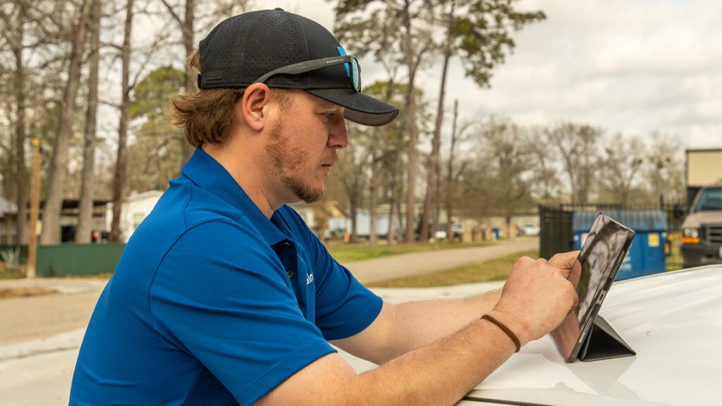 Roofing team member in a blue shirt and cap reviewing job details on a tablet outdoors