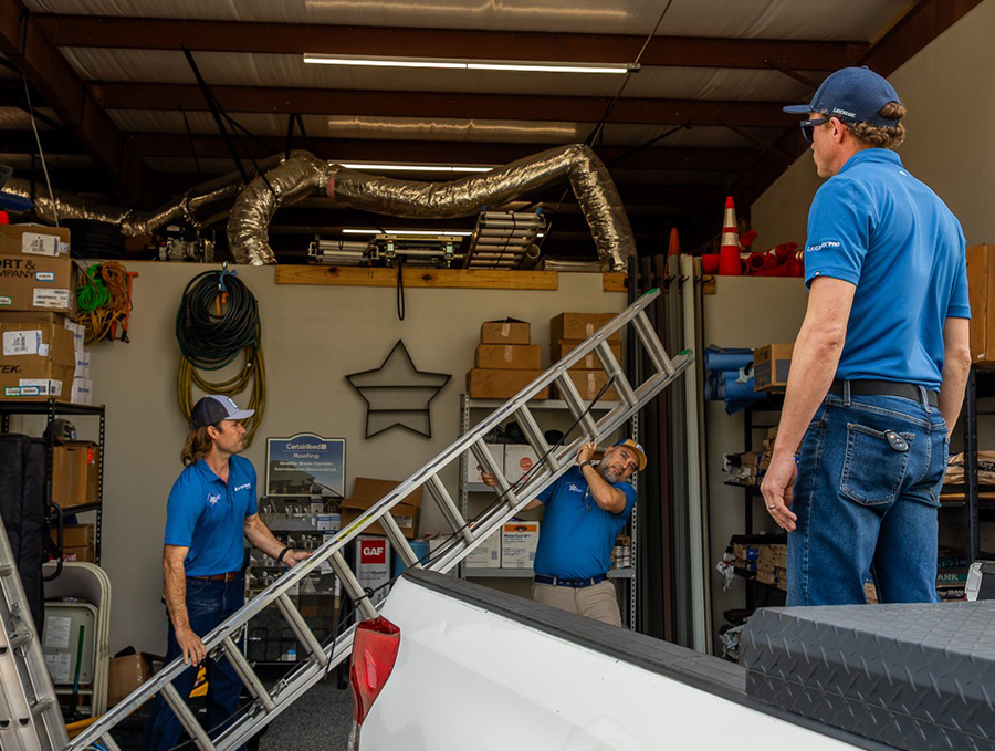 Roofing crew in blue shirts loading ladders and materials into a work truck inside the company warehouse