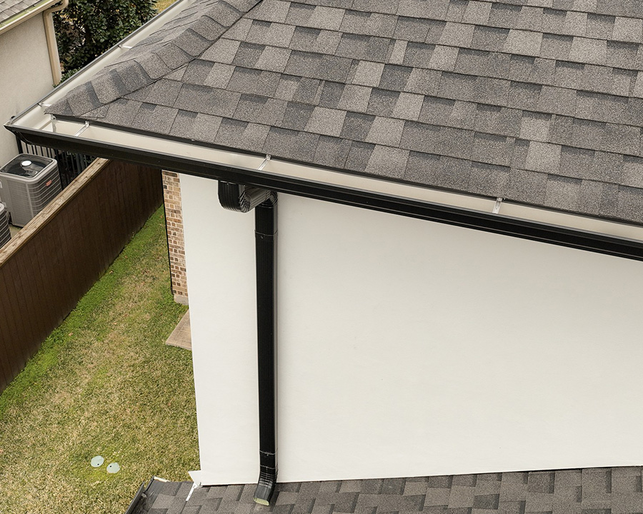 Dark shingle roof edge with a white gutter and downspout on a stucco home