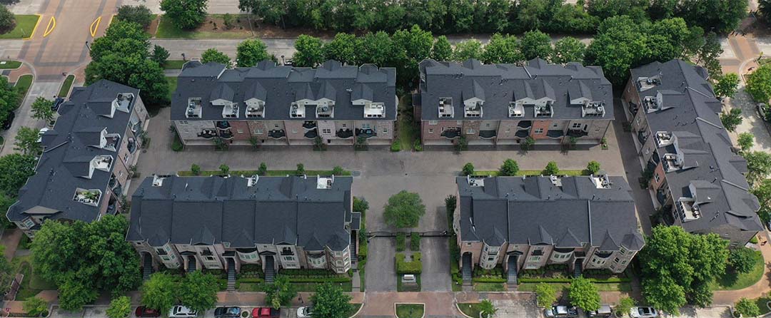 Aerial view of multi-family townhome buildings with dark gray shingle roofs in a wooded community