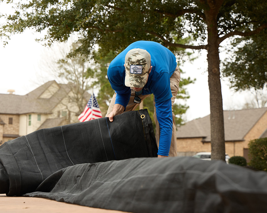 Roofer in a blue shirt unrolling a black protective tarp in a residential yard before starting a roof job