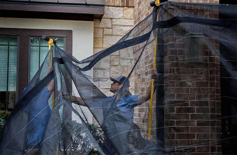 Worker installing black protective netting along a brick home exterior to catch debris during roofing work