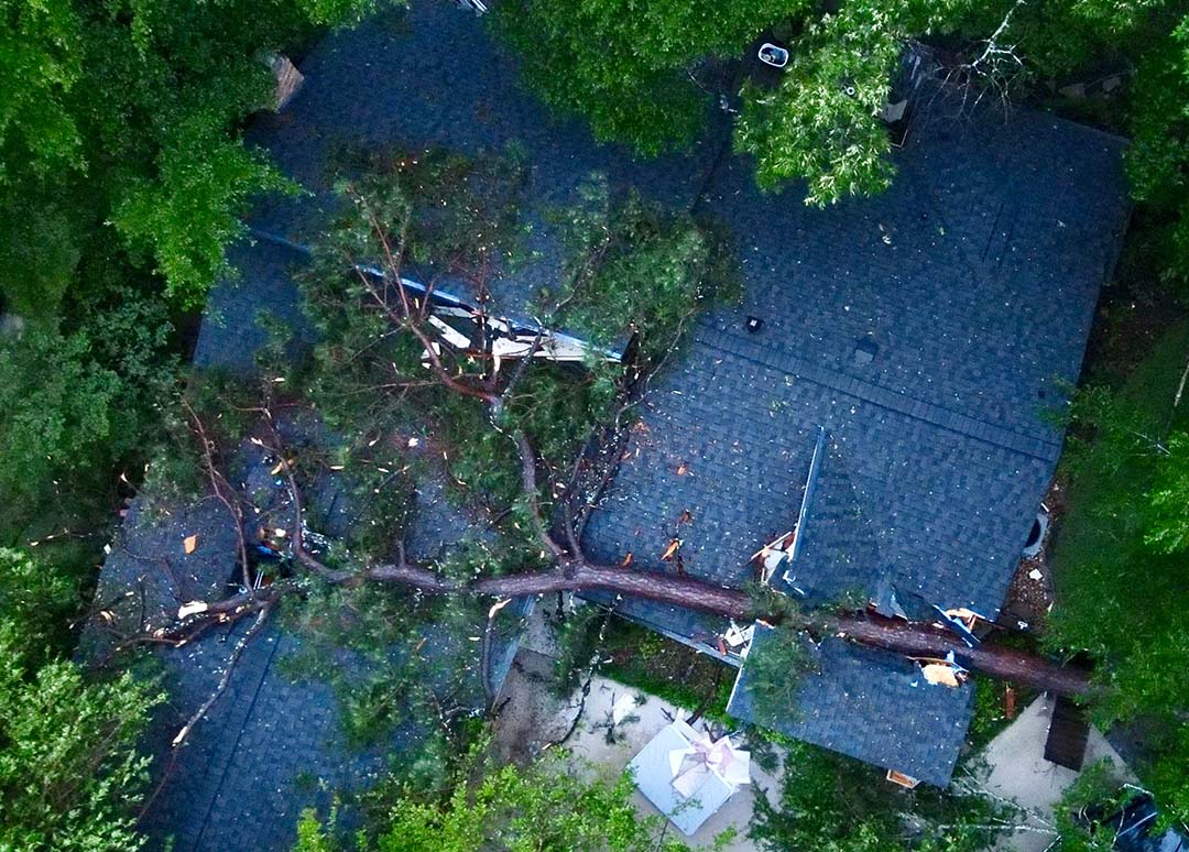 Aerial view of a large fallen tree lying across a home's blue-gray shingle roof after a storm