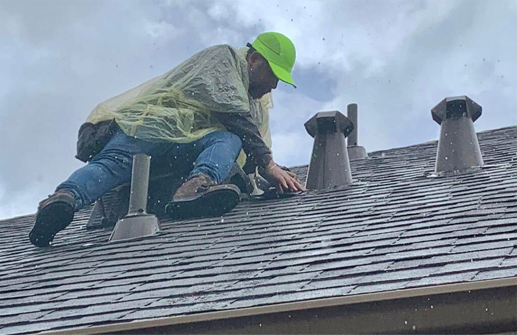 Roofer in a green hard hat and rain gear securing an emergency tarp on a shingle roof near vent pipes