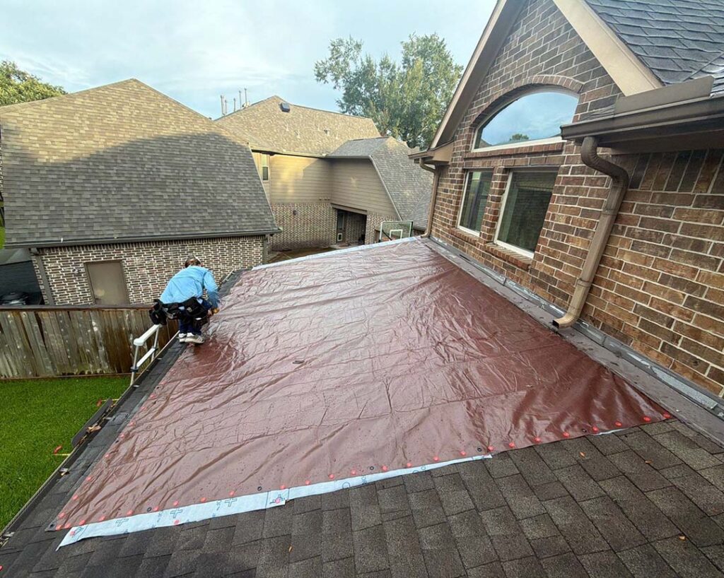 Brown emergency tarp secured over a damaged section of a shingle roof on a brick home