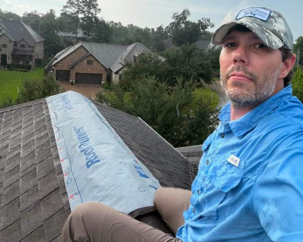 Roofer in a blue shirt and cap sitting on a roof beside a rolled emergency tarp